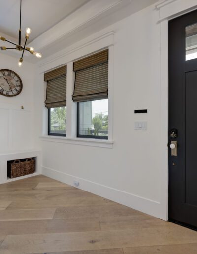 Bright entryway with a modern black door, wooden floor, and a bench with storage baskets underneath. A wall clock and a contemporary light fixture add style to the space.
