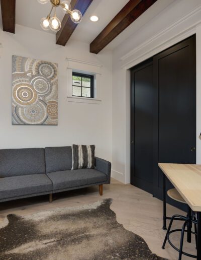 A modern room with a gray sofa, artwork on the wall, two small windows, a wooden desk, and plants. A dark rug lies on the wooden floor.