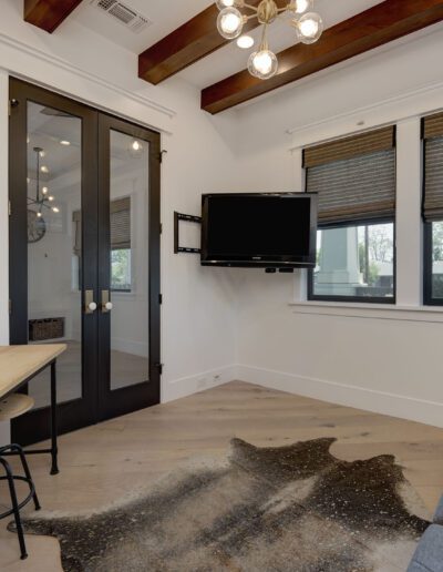 A modern home office with a desk, chair, lamp, and a wall-mounted TV. The room has wooden beams, a large window with blinds, a plant, and a sofa.
