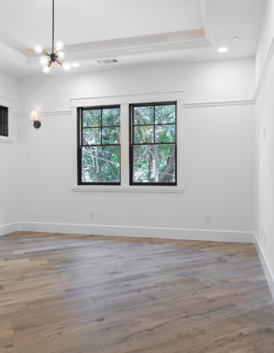 An empty room with white walls, wooden flooring, two large windows, and a modern ceiling light fixture.
