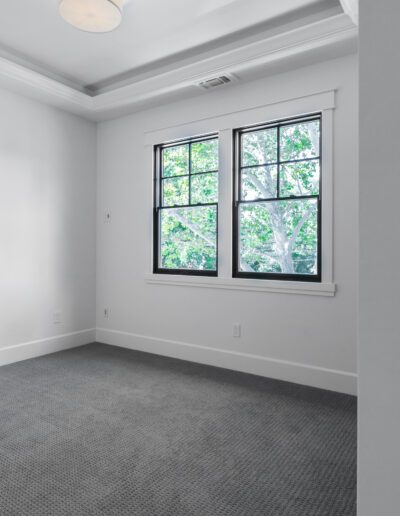 Empty room with gray carpet, white walls, a ceiling light, and a window showing green trees outside. A door with a white knob is partially open.