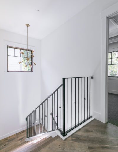 A stairwell with wooden steps, black metal railing, and a modern chandelier. A window and an open doorway are visible in the white-walled space.