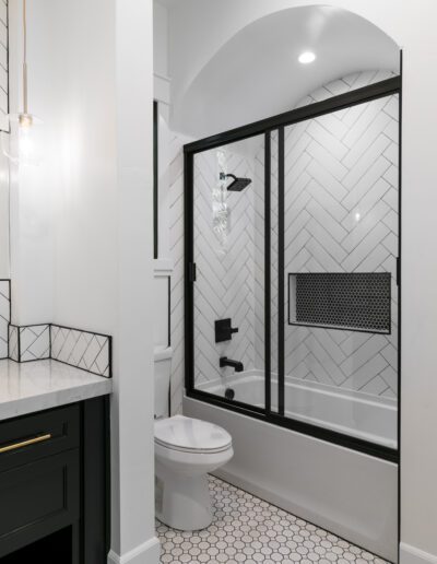 Modern bathroom with black and white tiles, featuring a bathtub with a glass door, black fixtures, hexagonal floor tiles, and a dark vanity with a marble countertop.