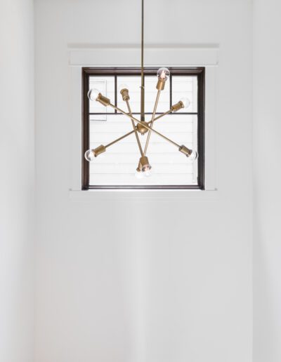 A brass sputnik chandelier with exposed bulbs hangs in front of a small rectangular window in a white room.