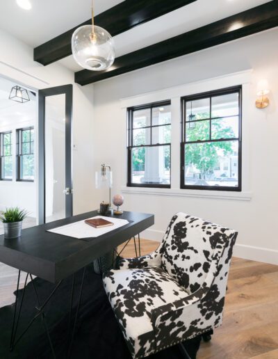 Modern office with wooden desk, cow-print chair, large windows, and ceiling beams. A globe, notepad, and plant decorate the desk.