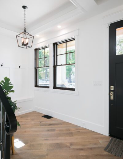 A bright, modern entryway with wooden flooring, a black front door, and stairs with a black railing. A large window and a plant are on the left side. A lantern-style light hangs above.