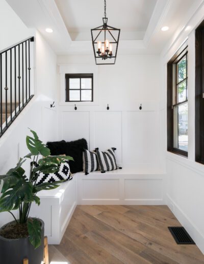 A bright entryway with wooden floors, a staircase with black railing, a bench with cushions, a potted plant, large windows, and a hanging light fixture.