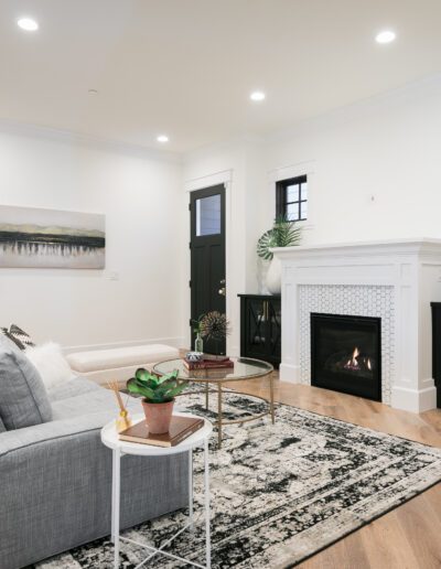 A modern living room with a gray sofa, a round glass coffee table, a fireplace, and a large indoor plant. The walls are white, and the floor is wooden with a patterned rug.