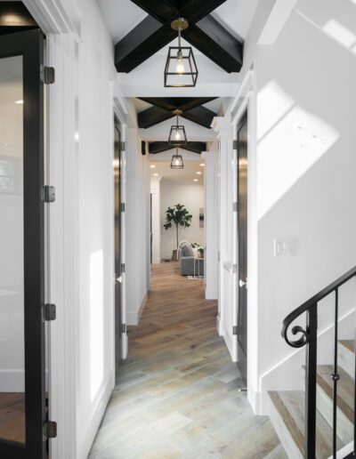 A hallway with wooden flooring, white walls, and black beam ceiling features pendant lights. Doors lead to adjacent rooms, and a living area with a plant is visible at the end.