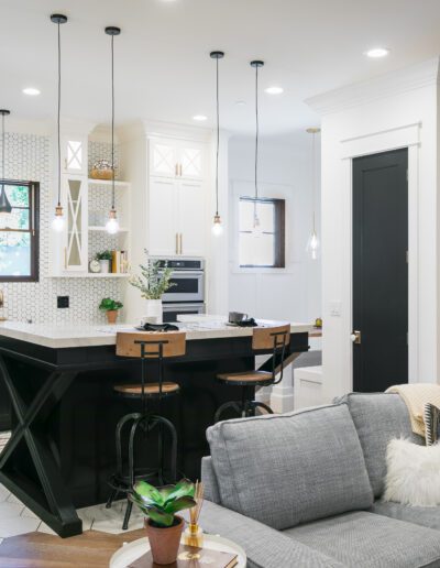 Modern kitchen and living area with black and white cabinetry, an island with stools, and a gray sofa. Decorative plants and light fixtures complement the space.
