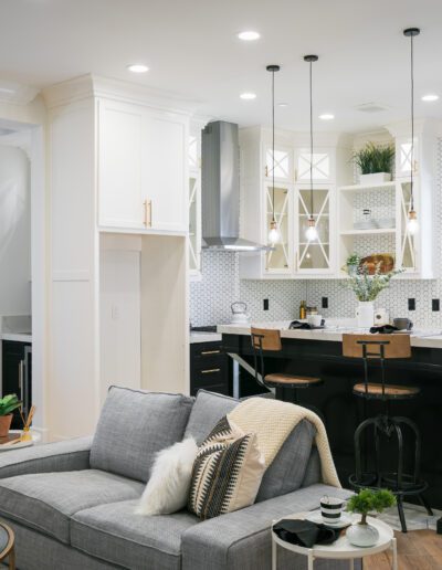Open-concept kitchen and living area with a modern design, featuring a gray sofa, black island with stools, white cabinets, and decorative lighting fixtures.