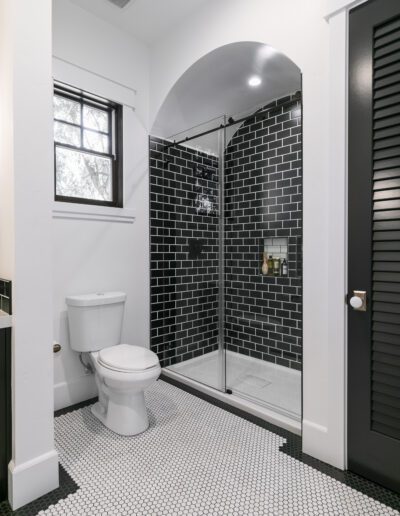 Bathroom with black tile shower, white walls, and hexagonal floor tiles. There is a toilet beside the shower and a window above.