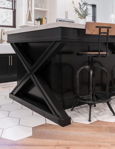 Modern kitchen with black island, hexagonal white floor tiles transitioning to wood, and high-back bar stools.