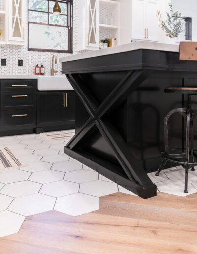 Modern kitchen with black island featuring X-shaped support, hexagonal white tile floor, and wooden flooring. White cabinets and gold accents are in the background.
