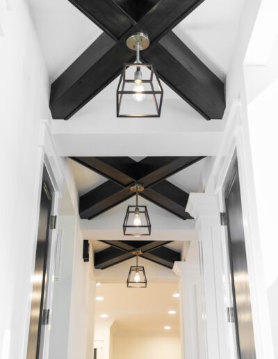 Hallway with black ceiling beams, geometric pendant lights, and white walls, leading to a room with a plant visible in the background.