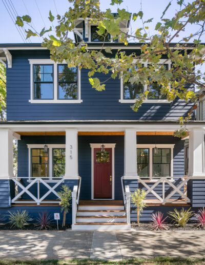 Two-story blue house with white trim, a red front door, and a covered porch. Surrounded by trees and small shrubs, with a sidewalk in front.