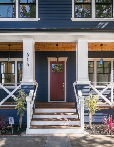 Front view of a blue house with a red door, white trim, and a porch with steps. House number 315 is visible. Plants are on either side of the steps.