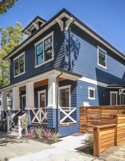 Blue two-story house with white trim, front porch, and a wooden fence. Surrounded by trees, with a clear blue sky above.