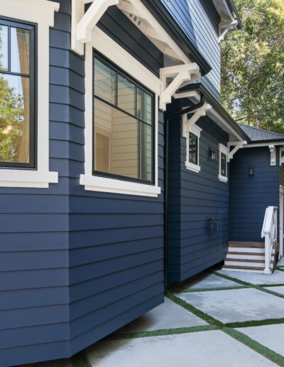 Side view of a modern blue house with white trim, featuring large windows and concrete walkway, adjacent to a wooden fence.