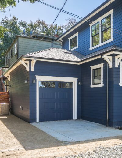 A blue house with white trim features a single-car garage. There's a gravel driveway and a wooden fence next to the garage. Tall trees are on the left side of the property.