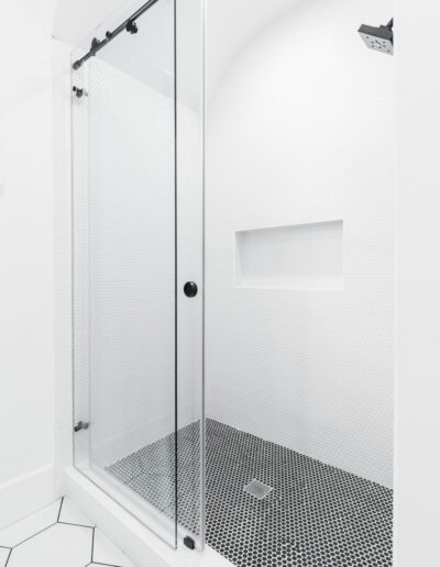 Minimalist shower with clear glass sliding door, white textured walls, built-in niche, and black hexagonal floor tiles.