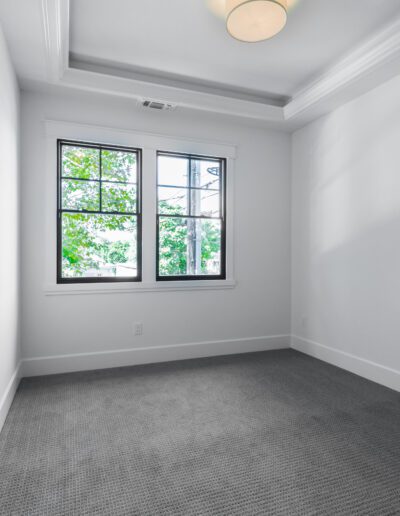 An empty room with gray carpet, white walls, a tray ceiling with a light fixture, and a window with a view of green trees.