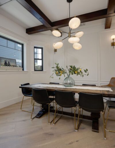 A modern dining room with a wooden table, eight black chairs, and a vase with flowers. A cluster of round pendant lights hangs above. Wood beam ceiling and large window in view.