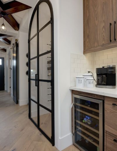 A modern kitchen with a coffee maker, dual faucets, a wine cooler, wooden cabinets, and a glass door. The hallway features exposed beams and leads to a dark front door.