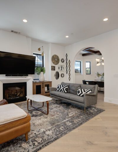 A modern living room with a gray sofa, brown leather chair, fireplace, large TV, and decorative rug. The room opens into a dining area with a small table and chairs.