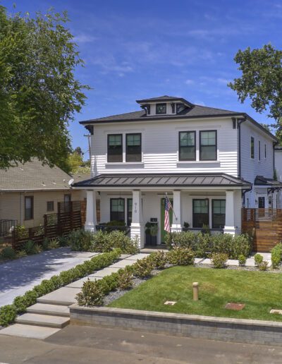 Two-story white house with black trim, surrounded by trees and greenery. Features a front porch, manicured lawn, and driveway. Clear blue sky in the background.