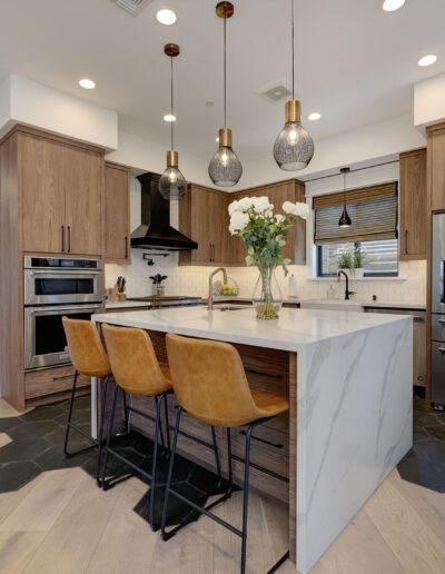 Modern kitchen with wood cabinets, stainless steel appliances, and a marble island. Three pendant lights hang above the island with three brown bar stools. Vase of flowers centered on island.