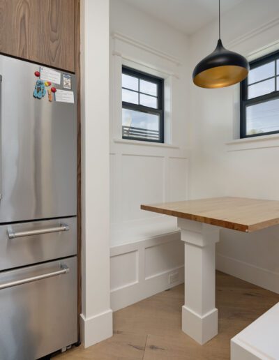 Modern kitchen nook with a stainless steel refrigerator, wooden cabinet, small wooden table, white bench seating, and two windows. Black pendant light hangs above the table.