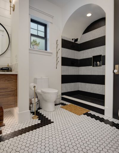 Modern bathroom with black and white hexagonal floor tiles, a wooden vanity, round mirror, and a shower with black and white striped tiles. A wicker basket holds folded towels.