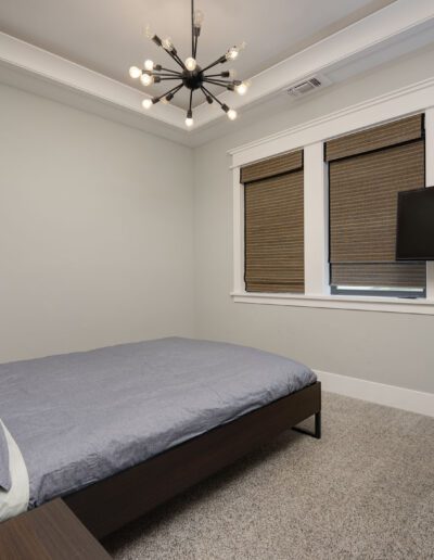 Bedroom with a single bed, modern chandelier, wall-mounted TV, and two windows with closed blinds. A small vase of flowers is on a side table.