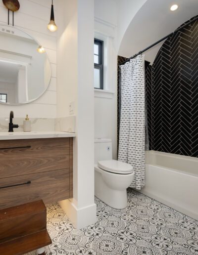 Modern bathroom with a wooden vanity, round mirror, and black herringbone tiled bathtub. Patterned floor tiles and a small stool are visible.