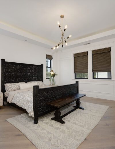 A bedroom with a dark wooden bed, beige bedding, a wooden bench at the foot, a nightstand, and a TV on the wall. Large windows with blinds and a modern light fixture are present.