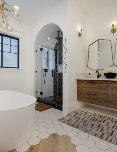 Modern bathroom with a freestanding tub, black-framed window, shower area, wood vanity with hexagonal mirrors, and geometric floor tiles.