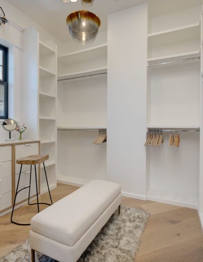 Spacious walk-in closet with white shelves, drawers, hanging rods, a small stool, a bench, and a ceiling light fixture. A large window illuminates the room.