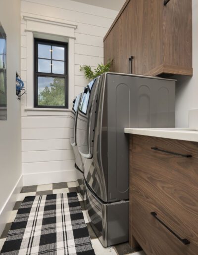 A narrow laundry room with a washer and dryer, wooden cabinets, a black faucet, and a black-and-white checkered rug. A window above the appliances provides natural light.