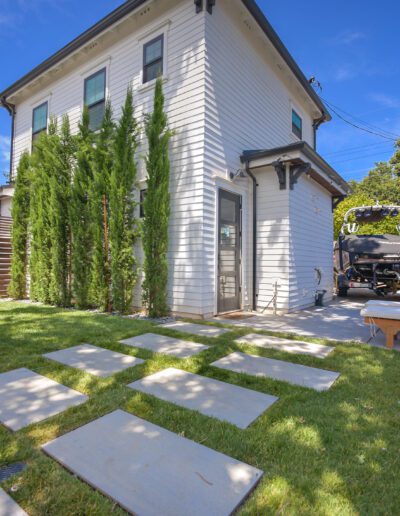 Backyard view with grass, stepping stones, and tall shrubs leading to a white house. A lounge chair and barbecue grill are on a patio, with a wooden fence surrounding the area.