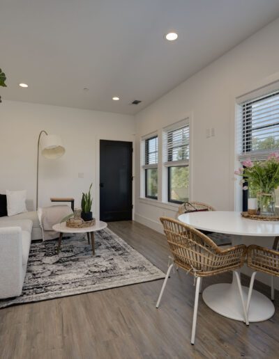 Modern living room with a white sofa, black and white rug, round coffee table, potted plants, and a small dining area with wicker chairs near large windows.