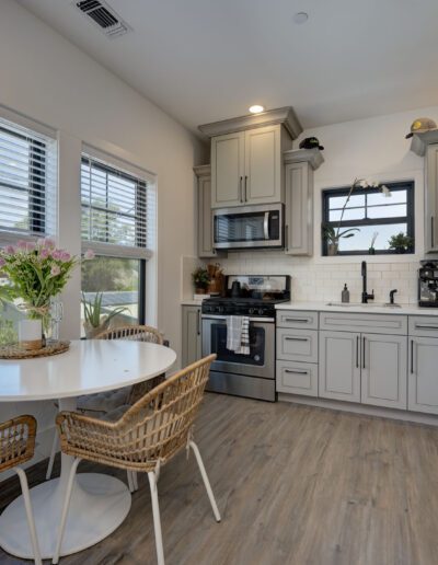 A modern kitchen with gray cabinets, stainless steel appliances, and a small dining area with a round table and wicker chairs. A plant and flowers add a touch of green.
