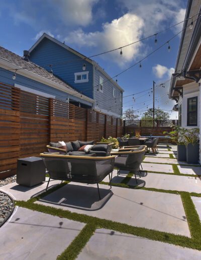 A modern backyard with outdoor seating, potted plants, string lights, and a stone path with grass. Wood fencing separates the space from neighboring houses.