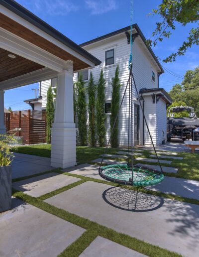 Modern backyard with a swing, lawn seating, potted plants, and a concrete pathway. White two-story house and wooden fencing in the background under a clear blue sky.