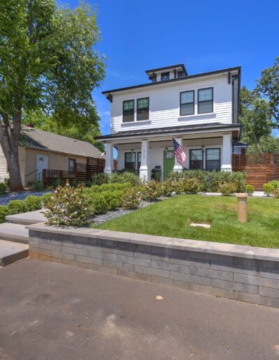 A two-story white house with a front porch, manicured lawn, and a U.S. flag stands under a clear blue sky, surrounded by trees and neighboring homes.
