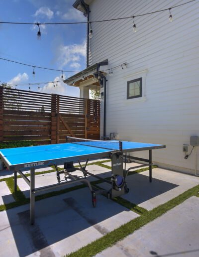 A blue ping pong table stands on a patio with string lights above, surrounded by white walls and wooden fencing.
