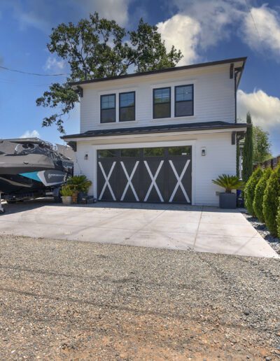 A two-story modern house with a boat parked on a paved driveway. The house has two garage doors and is surrounded by a wooden fence and trees under a clear blue sky.