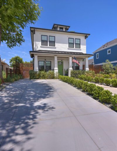 Two-story white house with an American flag, flanked by a beige house on the left and a blue house on the right. Concrete driveway and well-maintained shrubs lead to the entrance.