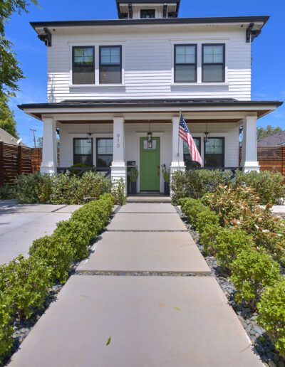 A two-story white house with a green door and American flag, surrounded by greenery and a concrete walkway, on a bright, sunny day.