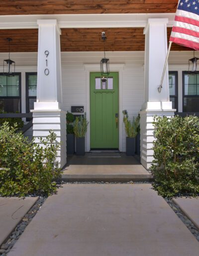 Front porch of a house with a green door and American flag, surrounded by greenery. House number 910 is displayed on the wall.
